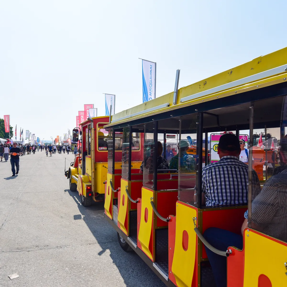 Besucher nutzen einen Bummelzug auf dem Gelände mit Ausstellern und Messeständen im Hintergrund.